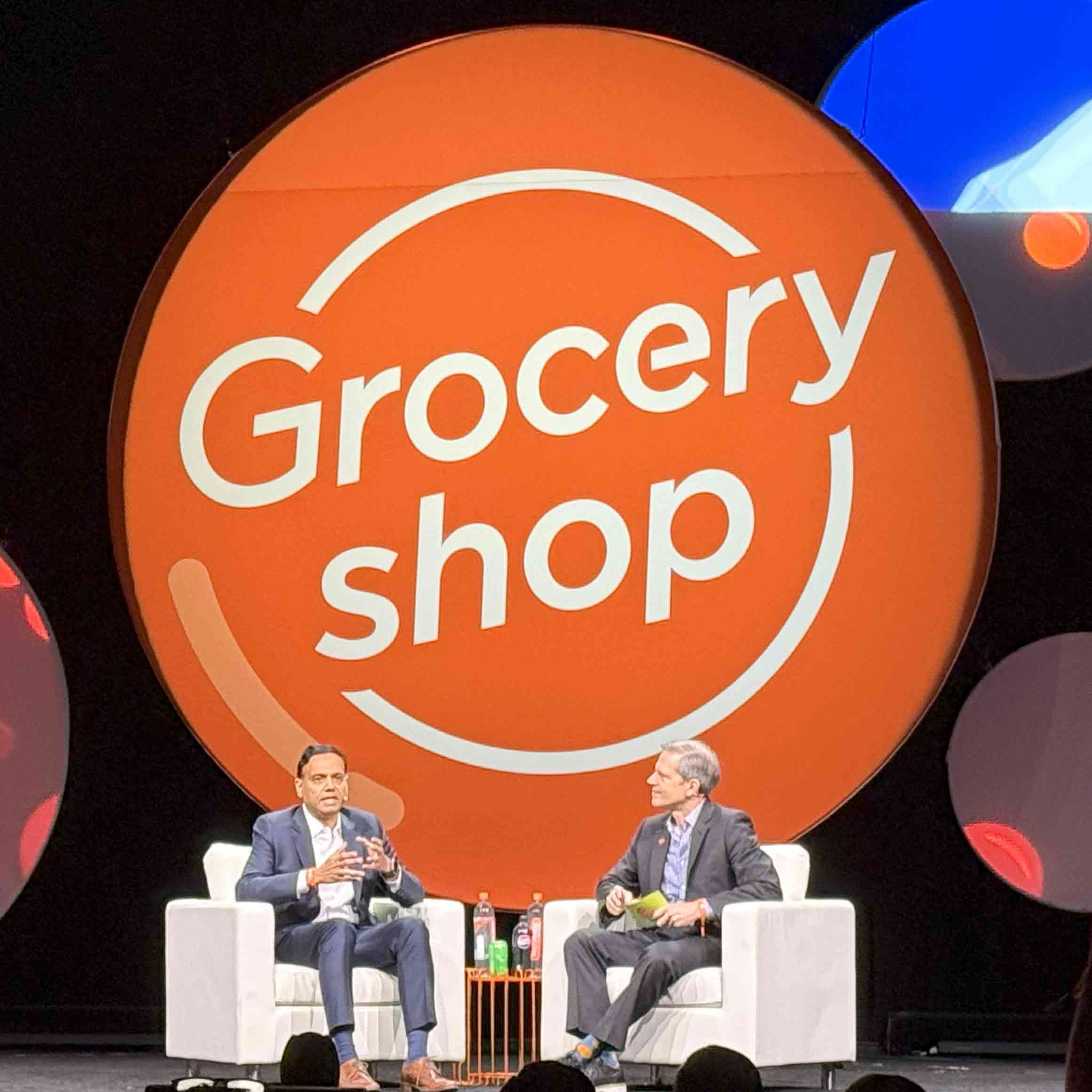 Two men speak in large chairs in front of a background from Grocery Shop